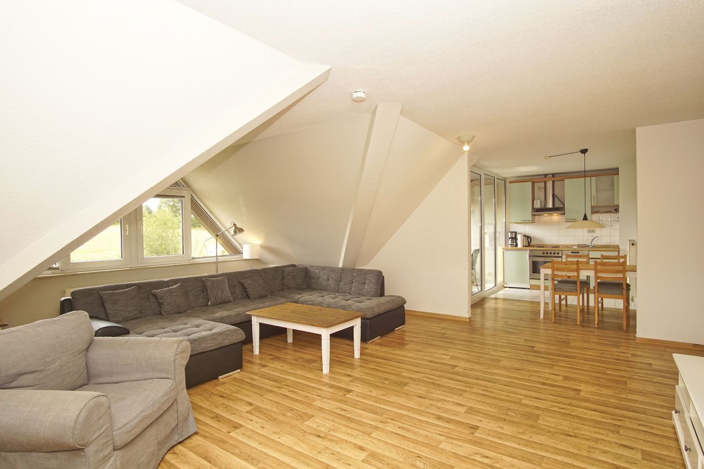 Un appartement moderne de style « loft » avec un plafond en pente, un canapé d’angle, une petite table en bois et un coin cuisine avec un ensemble de salle à manger.
