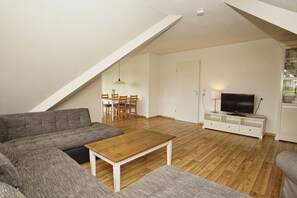 A modern living room with a wooden coffee table, grey sofas, a TV on a white cabinet, and a dining area with wooden chairs and a table. - Close to the beach with a balcony; access to the pool and sauna - Strandhaus Mönchgut FeWo21 (Middelhagen)