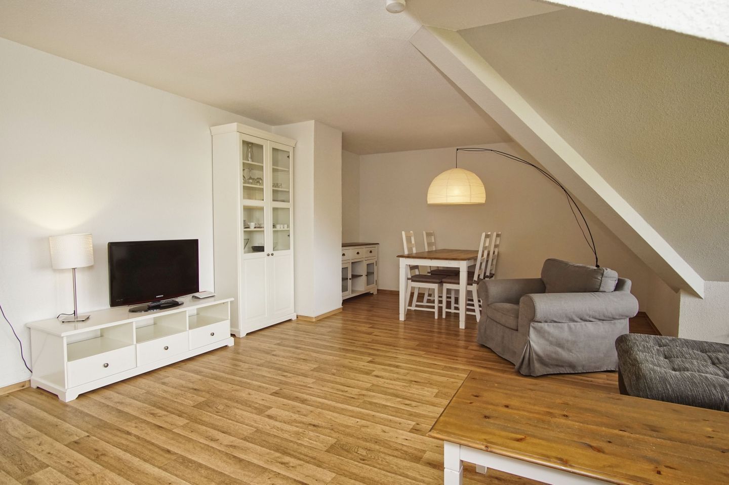 Ein modernes Wohnzimmer mit Fernseher, weißem Schrank, Esstisch und grauem Sofa.