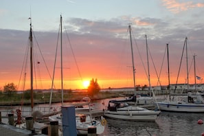 A marina with multiple sailboats docked, a sunset in the background, and a calm water surface.