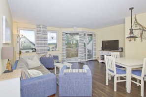 A living room with a sofa, armchairs, a dining table with chairs, a TV, and a balcony with curtains.