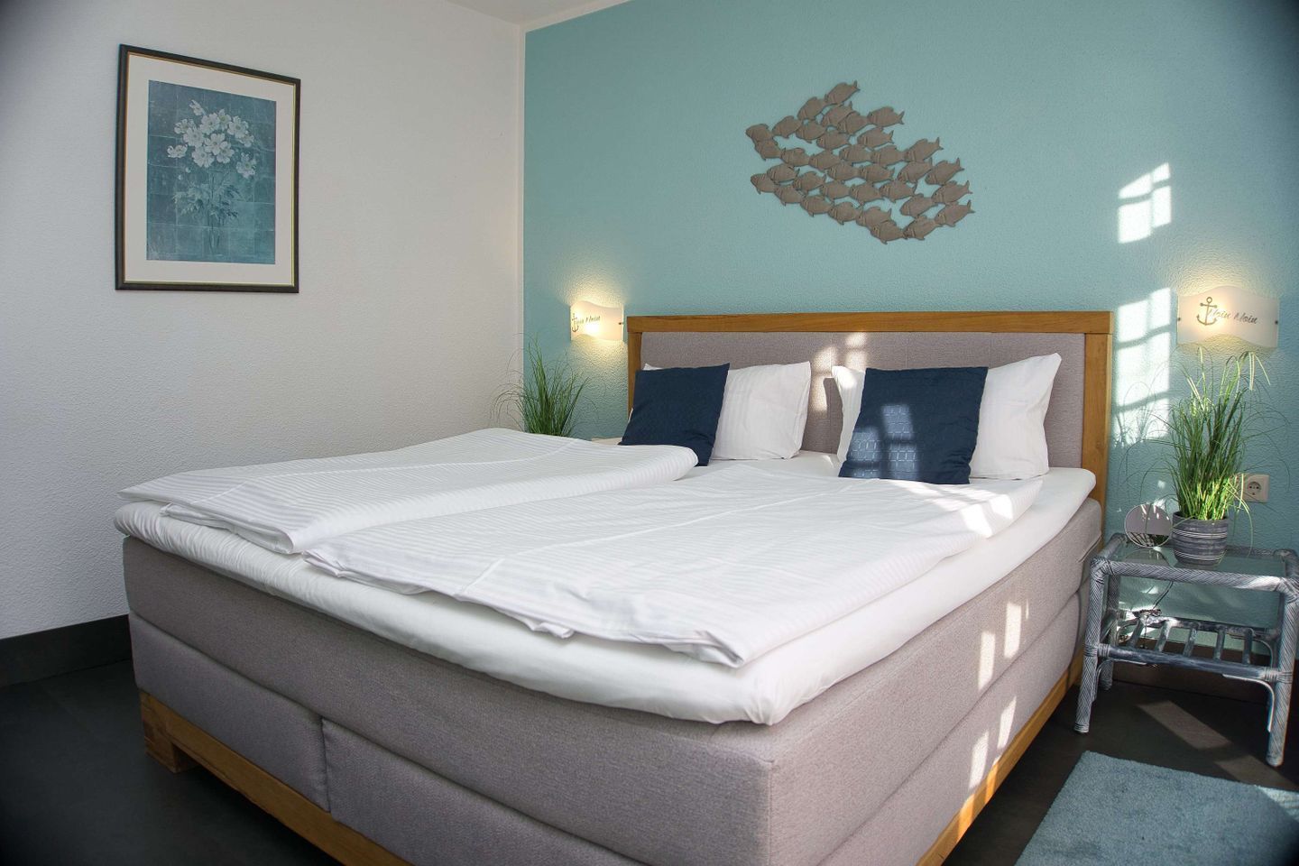 A neatly made bed with white linens, a light blue wall, a decorative wall piece, and a small table with a plant.