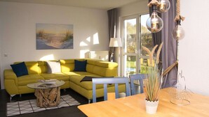 A yellow sofa with blue cushions, a round glass coffee table, a wooden side table, and a wooden dining table with a white vase and a metal sculpture.
