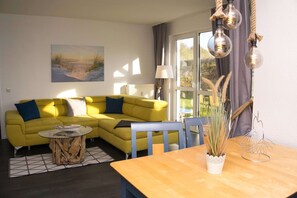 Un canapé jaune avec des coussins bleus, une table basse ronde en verre, une table d’appoint en bois et une table à manger en bois agrémentée d’un vase blanc et d’une sculpture en métal.