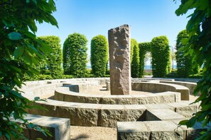 Ein Steinsockel, umgeben von einem kreisförmigen Steinweg, mit ordentlich geschnittenen Hecken und Bäumen im Hintergrund.