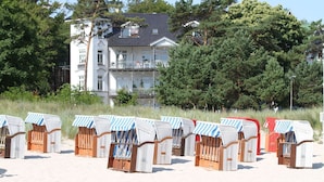 Strandkörbe mit gestreiften Sonnendächern auf einem sandigen Strand, mit einem mehrstöckigen Gebäude und Bäumen im Hintergrund.