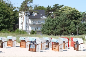 Strandkörbe mit gestreiften Sonnendächern auf einem sandigen Strand, mit einem mehrstöckigen Gebäude und Bäumen im Hintergrund.