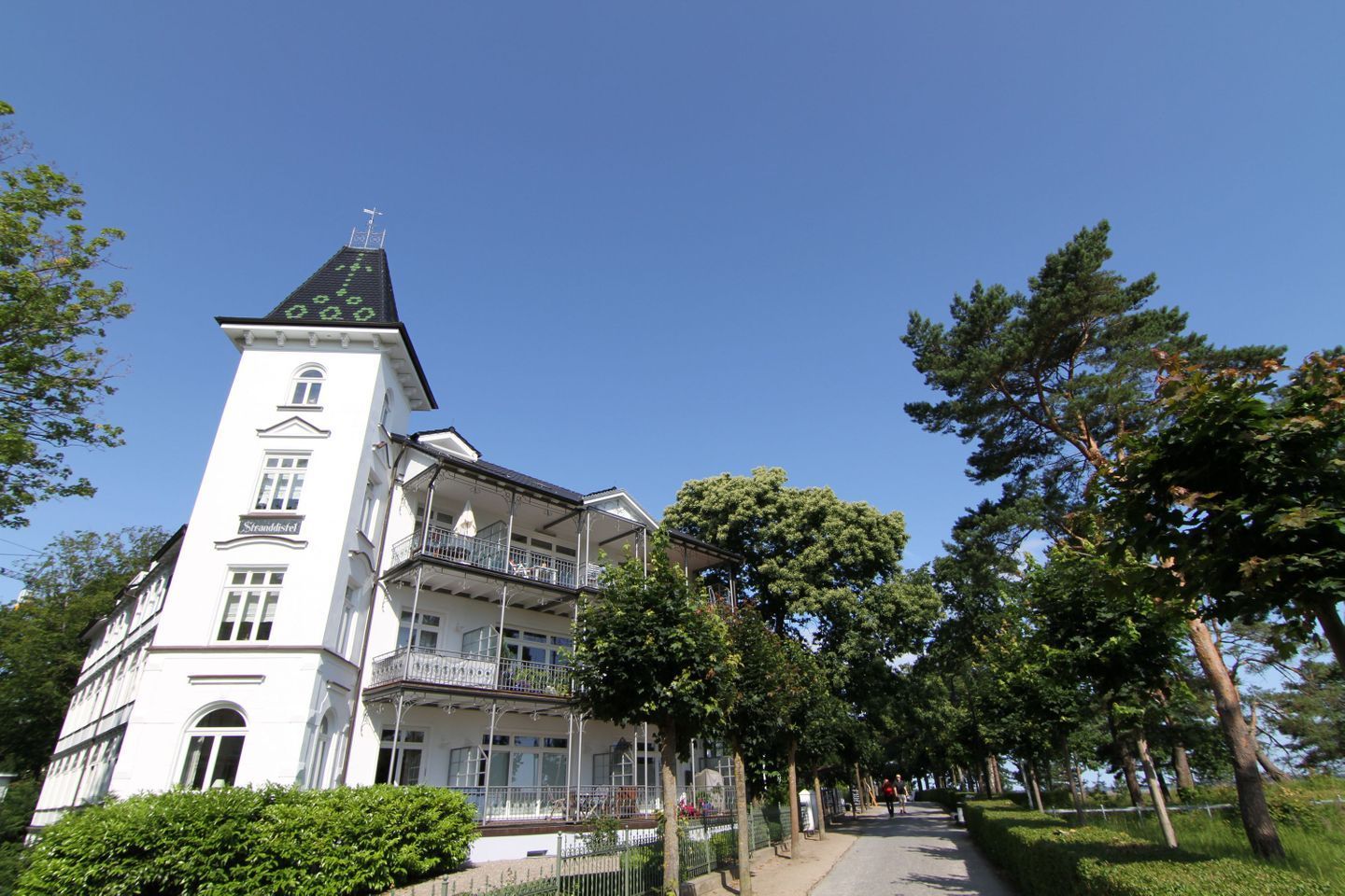 Un bâtiment blanc doté d’une tour, de balcons et d’un chemin bordé d’arbres.