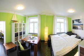 Una habitación de hotel con una cama, un escritorio con una silla, una estantería y una ventana con cortinas.