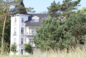 A white building with multiple balconies and a steeply pitched roof, surrounded by dense pine trees. - Beachfront vacation apartment in a classic seaside style - Villa Stranddistel FeWo 3.6 (Binz)