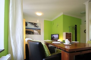 A room with a wooden dining table, a black leather chair, a white curtain, a painting of a beach, a television, and a green wall. - Beachfront vacation apartment in a classic seaside style - Villa Stranddistel FeWo 3.6 (Binz)
