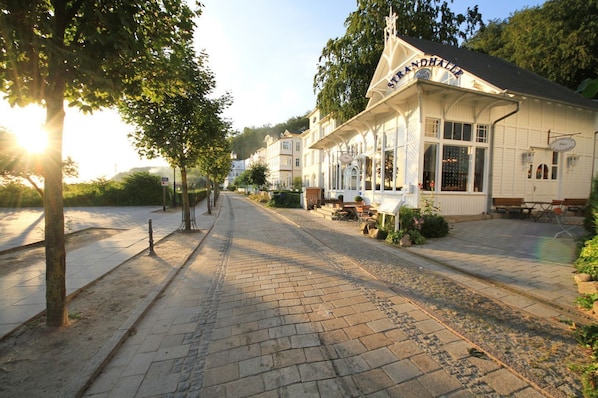 Un bâtiment sur lequel on peut lire l’inscription « Strandhalle », ainsi qu’une rue pavée bordée d’arbres et de bancs.