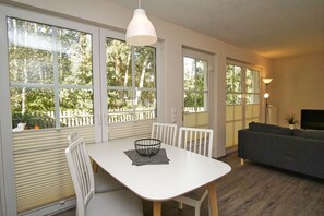 Una sala de estar moderna con una mesa de comedor blanca, sillas blancas, un sofá gris y ventanas amplias que dan a vista de árboles y una cerca.