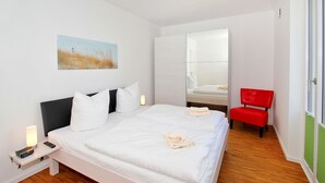 A modern hotel room with a large bed, a bedside table with a lamp, a red chair, and a painting on the wall. - Dünenresort Binz - beachfront, chic vacation apartment with balcony directly in Binz - (Binz)