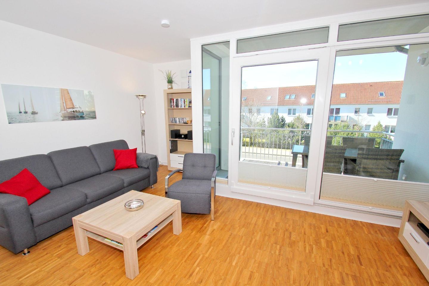 A modern living room with a grey sofa, a small armchair, and a wooden coffee table. There is a bookshelf with books and decorative items, and a large window offering a view of buildings and greenery.