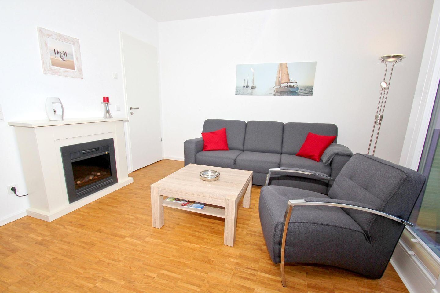 A living room with a fireplace, grey sofa with red cushions, a wooden coffee table, and a framed picture on the wall.