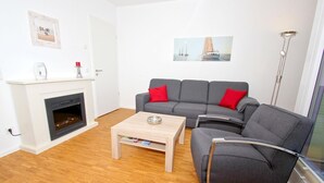 A living room with a fireplace, grey sofa with red cushions, a wooden coffee table, and a framed picture on the wall. - Dünenresort Binz - beachfront, chic vacation apartment with balcony directly in Binz - (Binz)