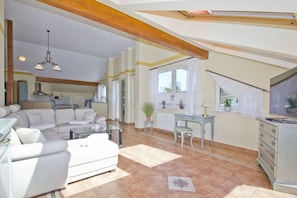 Ein geräumiges Wohnzimmer mit einer Couch, einem Glastisch und einem Schreibtisch mit Stuhl. Im Hintergrund befindet sich eine Küche, ein Fenster mit Vorhängen und eine Decke mit sichtbaren Holzbalken.