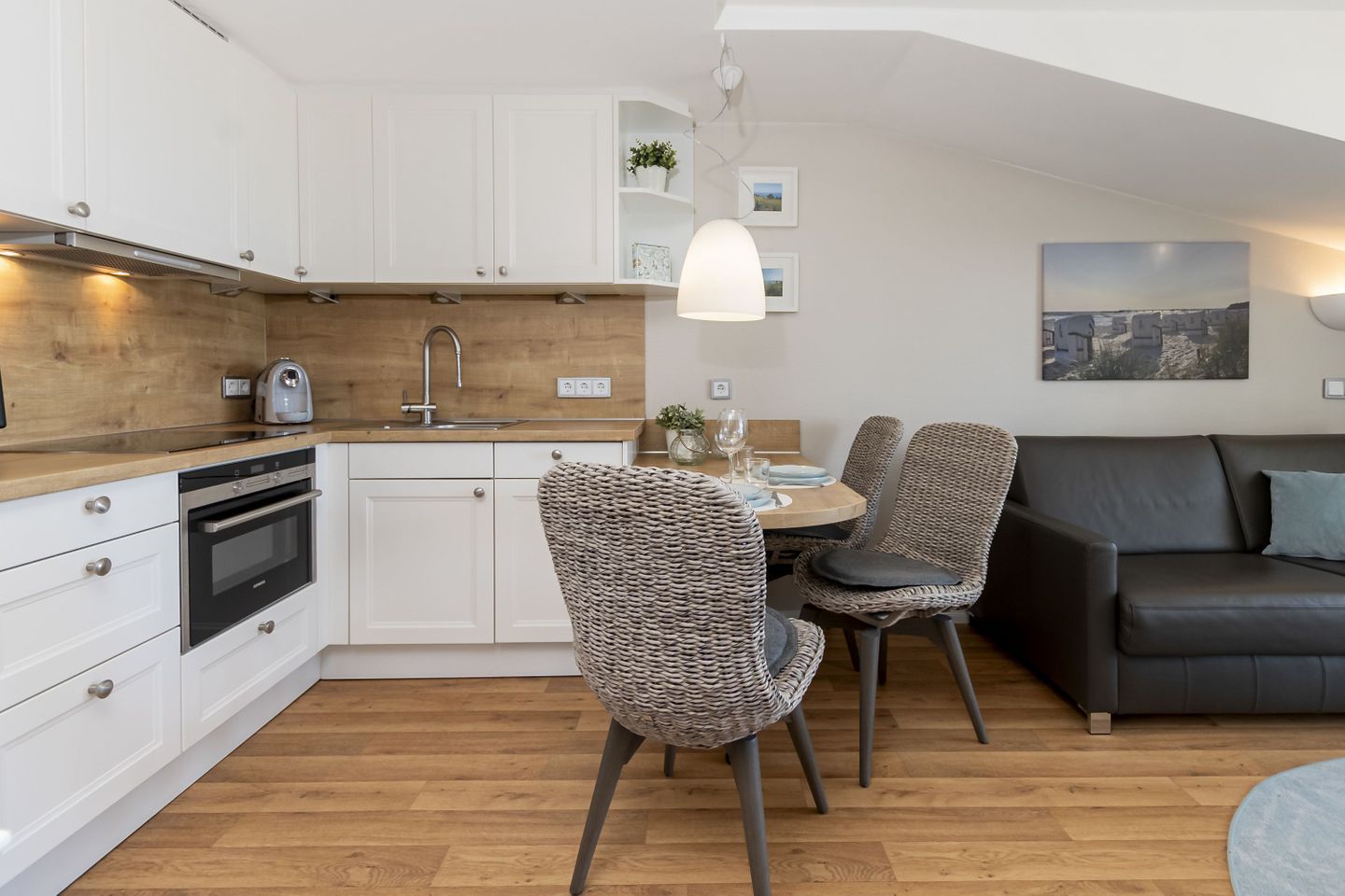 Een moderne keuken met witte kasten, een ingebouwde oven en een houten aanrecht. Er is een zithoek met rieten stoelen en een zwarte bank.