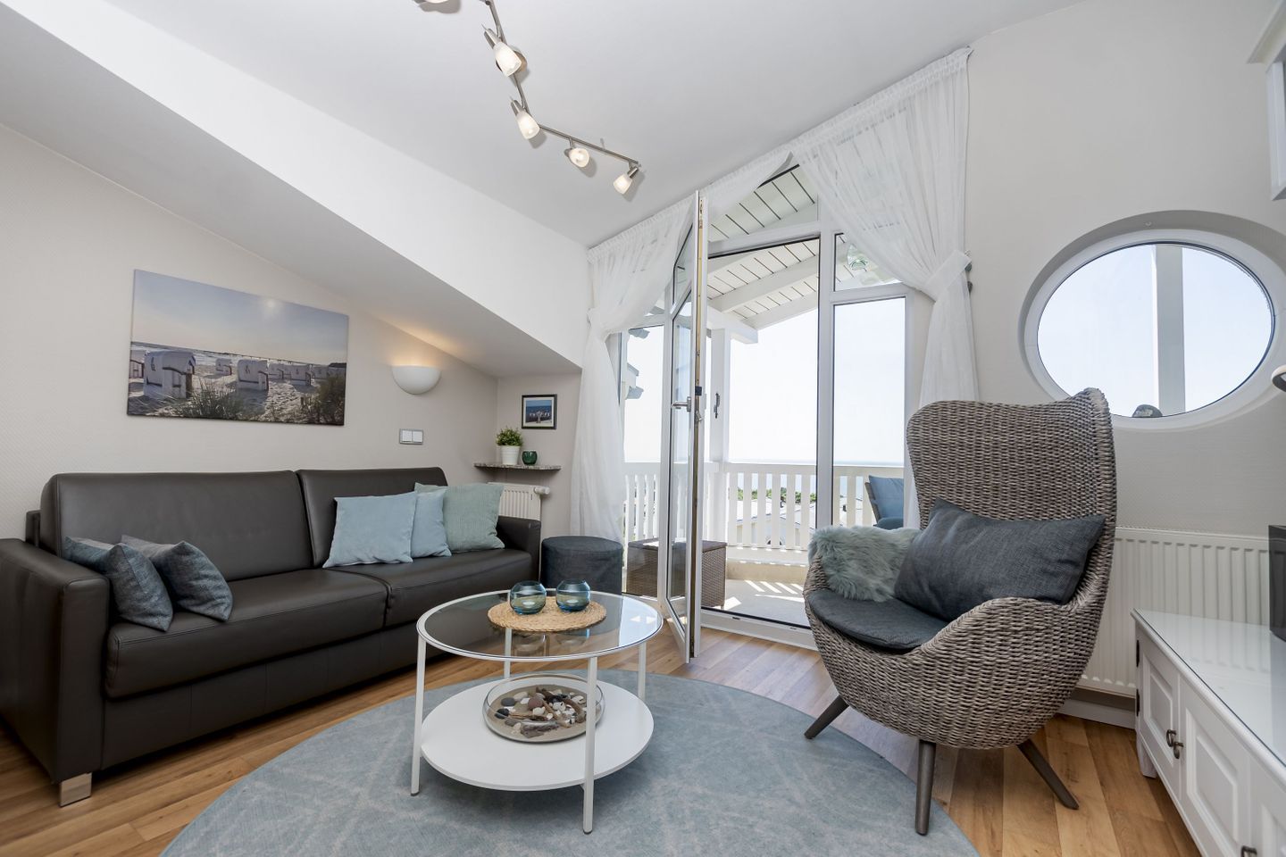 A modern living room with a sofa, a round wicker chair, a glass coffee table, and a balcony with a view.