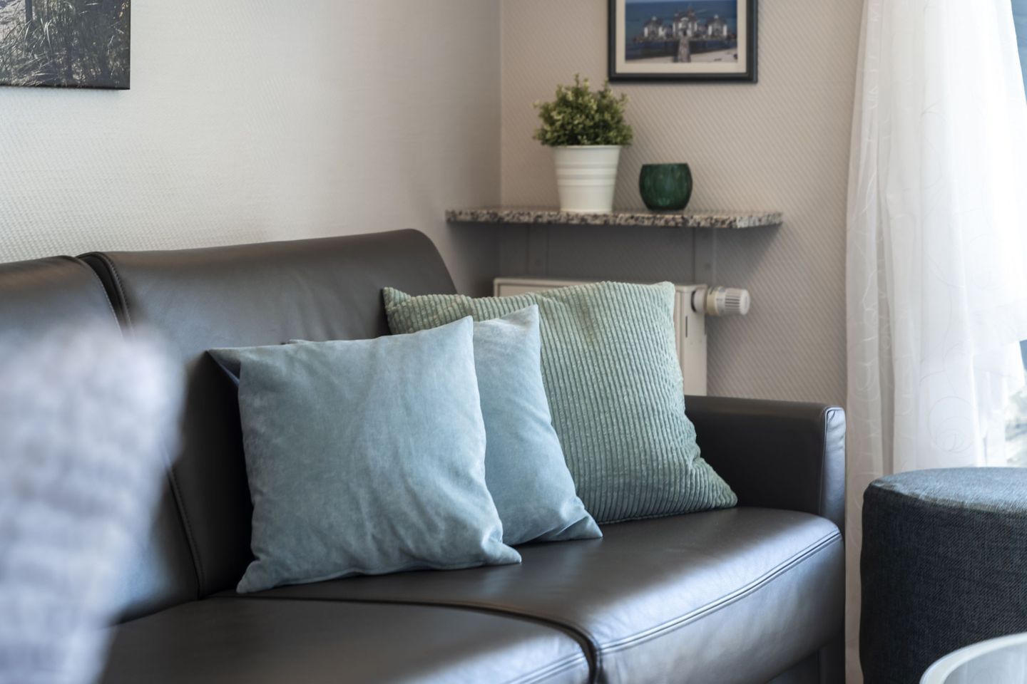 En soffa klädd i läder med två kuddar, en liten hylla med en växt och en dekorativ prydnad samt en inramad tavla på väggen.