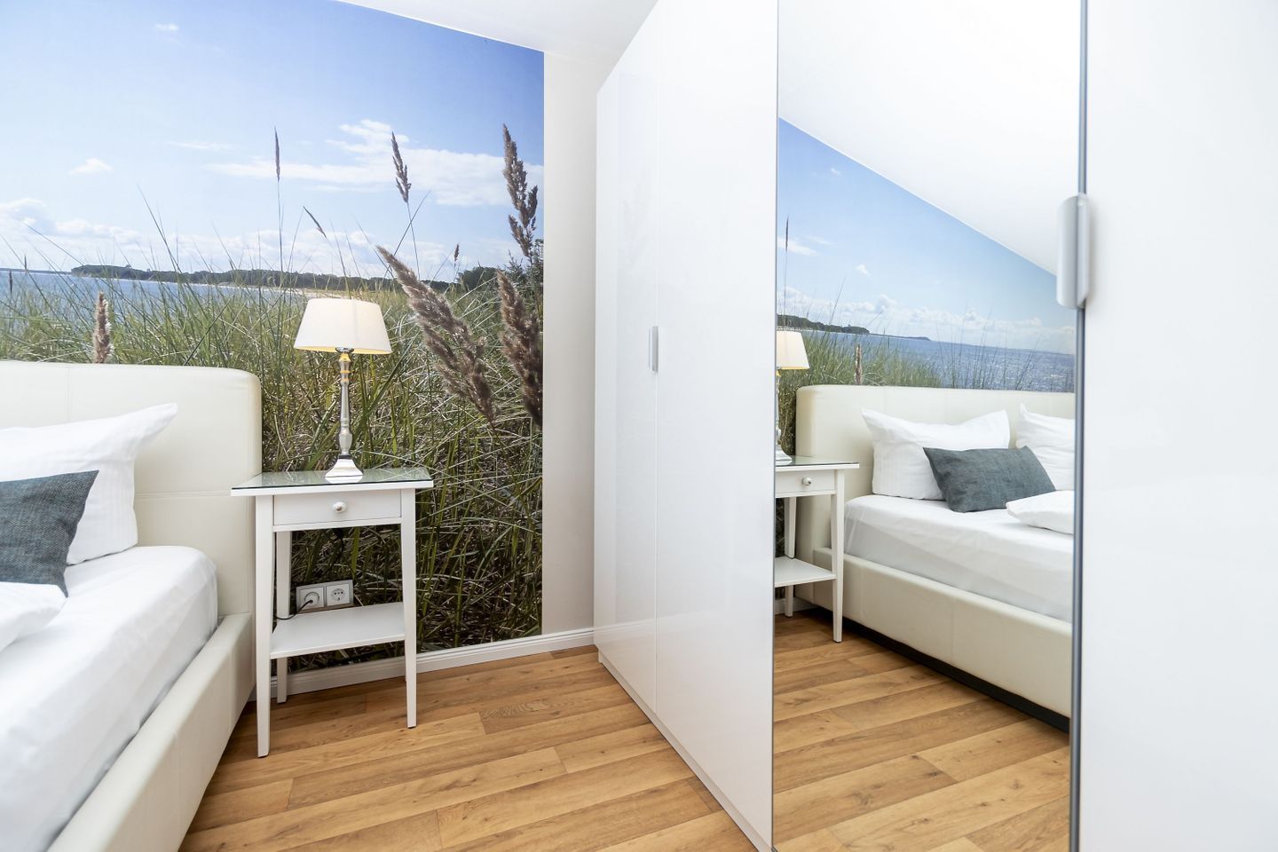 A bedroom with a white bed, a bedside table with a lamp, and a view of a beach through a large window.