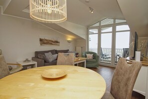 A spacious living area with a round wooden table, a green armchair, and a sofa with a striped pillow. A large window offers a view of the outdoors.