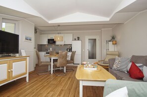 A modern living room with a flat-screen TV, a wooden coffee table, a sofa with cushions, and a dining area with chairs and a kitchen in the background.