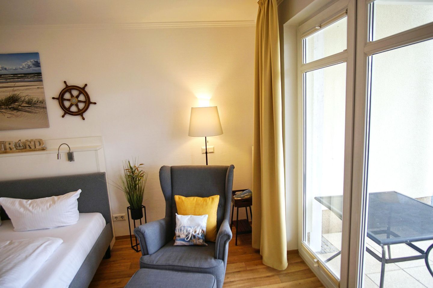 A hotel room with a bed, a chair with a yellow pillow, a lamp, a plant, and a view of the outdoors through a large window.