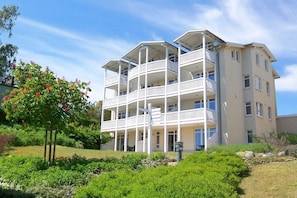A multi-story building with balconies, surrounded by greenery and a clear sky.