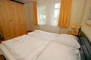 A double bed with white linens, a wooden wardrobe, a window with yellow curtains, and a bedside table with a lamp. - Patio, free use of the AHOI adventure pool and sauna - Apartment 10 with ocean view - (Göhren)