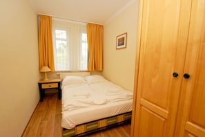 A small, single-bed room with a wooden floor, a nightstand, a lamp, and a window with yellow curtains.