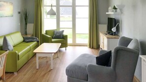 A living room with a green sofa, a grey armchair, a small white table, and a TV on a cabinet.