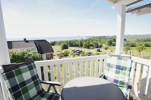 Ein Balkon mit weißer Brüstung, zwei Stühle mit Karokissen und Blick auf eine grüne Landschaft sowie Gebäude in der Ferne.