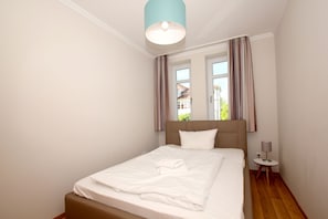 A neatly made bed with white linens and a brown headboard, a small bedside table with a lamp, and a window with curtains. - With a view of the sea and a balcony, access to the pool and sauna - Sea View Apartment 26 - (Göhren)