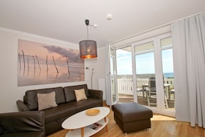 A modern living room with a large window, a brown leather sofa, a white coffee table, and a pendant light. - With a view of the sea and a balcony, access to the pool and sauna - Sea View Apartment 26 - (Göhren)