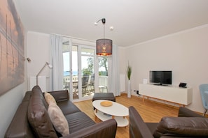 A modern living room with a sofa set, a coffee table, a TV on a white cabinet, and a view of the outdoors through a glass door. - With a view of the sea and a balcony, access to the pool and sauna - Sea View Apartment 26 - (Göhren)