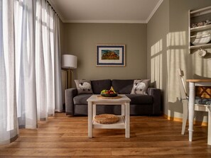 Uma sala de estar com um sofá cinzento, uma mesa pequena branca com uma cesta de palha, e um quadro pendurado na parede.