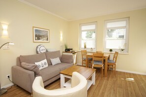 A cozy living room with a beige sofa, a wooden coffee table, and a dining table with chairs. There are two windows with blinds, a lamp, and a framed picture on the wall.
