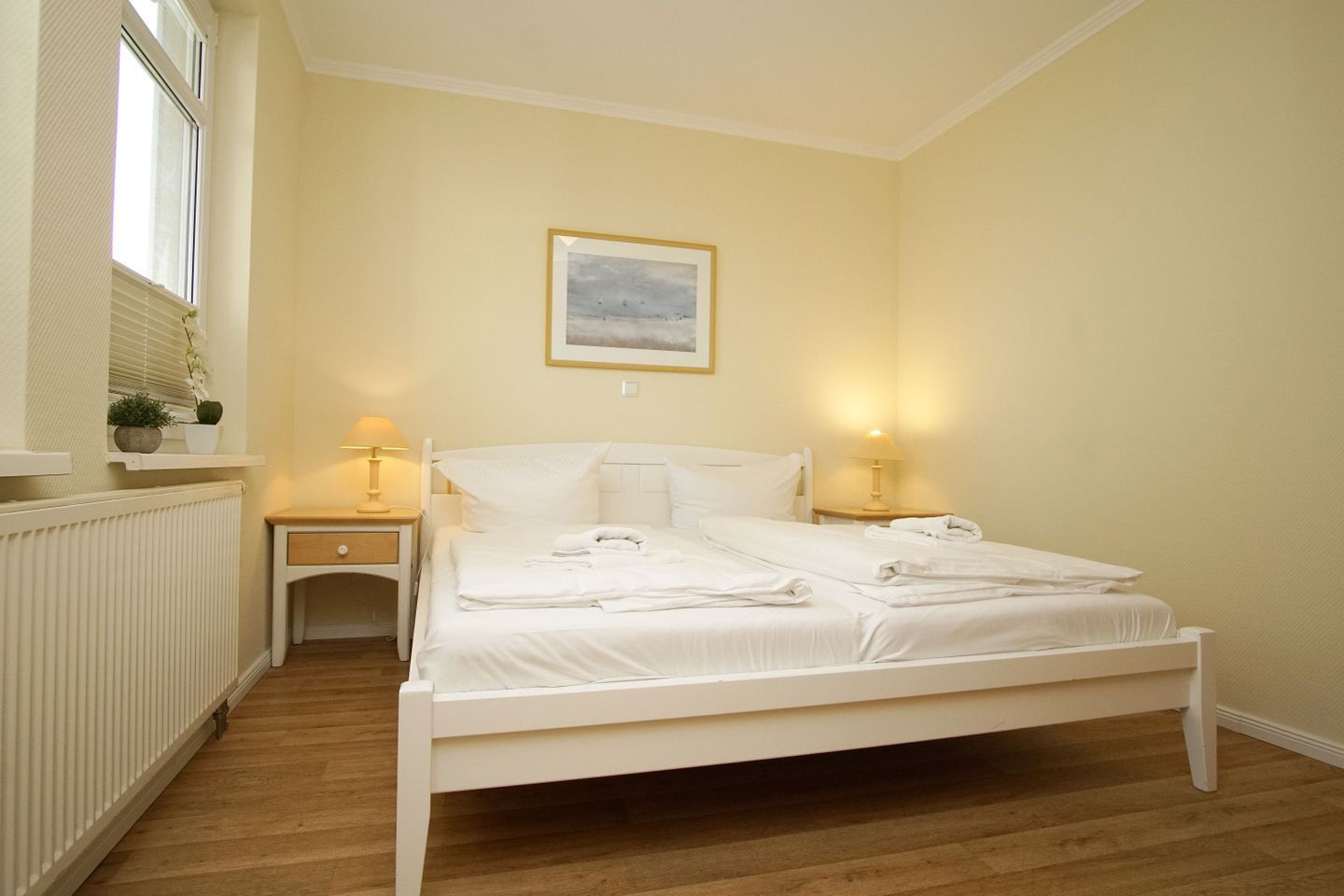 Un dormitorio con cama blanca, dos mesitas de noche con lámparas, una ventana con persianas y un radiador.