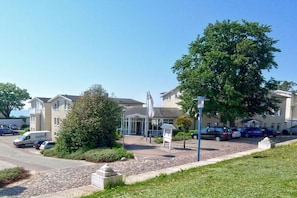A building with a parking lot, a large tree, and a statue in the foreground. - Patio, free use of the AHOI adventure pool and sauna - Apartment 44 with ocean view - (Göhren)