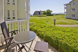 A patio with a round table and two chairs, a hedge, and a view of a house in the background.