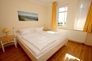A bedroom with a large bed, two bedside tables, a window with curtains, a radiator, and a painting on the wall.