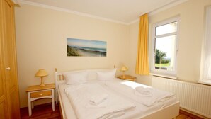 A neatly made bed with white linens and towels, a bedside table with a lamp, a window with curtains, and a painting on the wall.