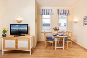 A cozy room with a TV on a wooden cabinet, a dining table with chairs, and two windows with striped valances.