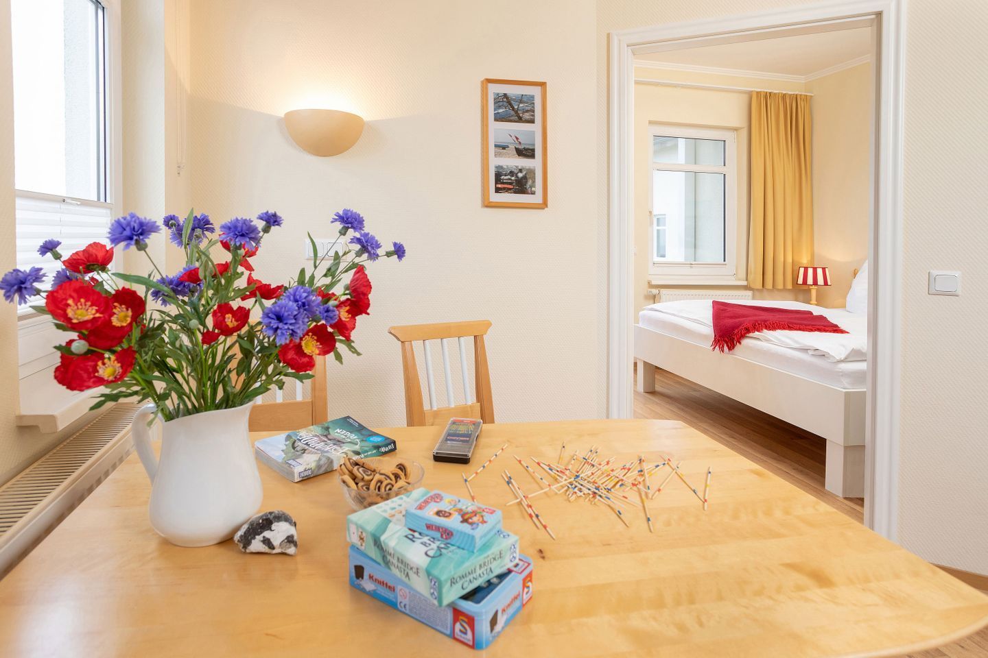 Une pièce avec une table à manger en bois, un vase de fleurs, un lit recouvert d’une couverture rouge et un tableau encadré accroché au mur.