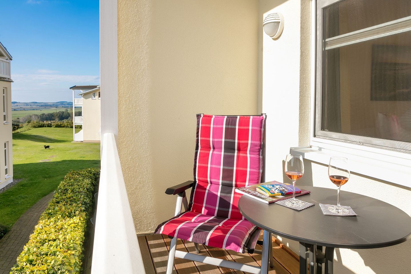 A balcony with a red and black striped chair, a small table with a glass of wine, and a book.