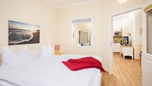 A hotel room with a bed, a painting of a beach, a mirror, and a kitchenette. - Balcony, free use of the AHOI adventure pool and sauna - Sea view apartment 45 - (Göhren)