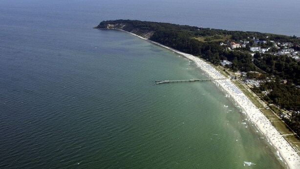 An aerial view of a coastal area with a long pier extending into the sea, a sandy beach, and a densely wooded peninsula. - Balcony, free use of the AHOI adventure pool and sauna - Sea view apartment 45 - (Göhren)