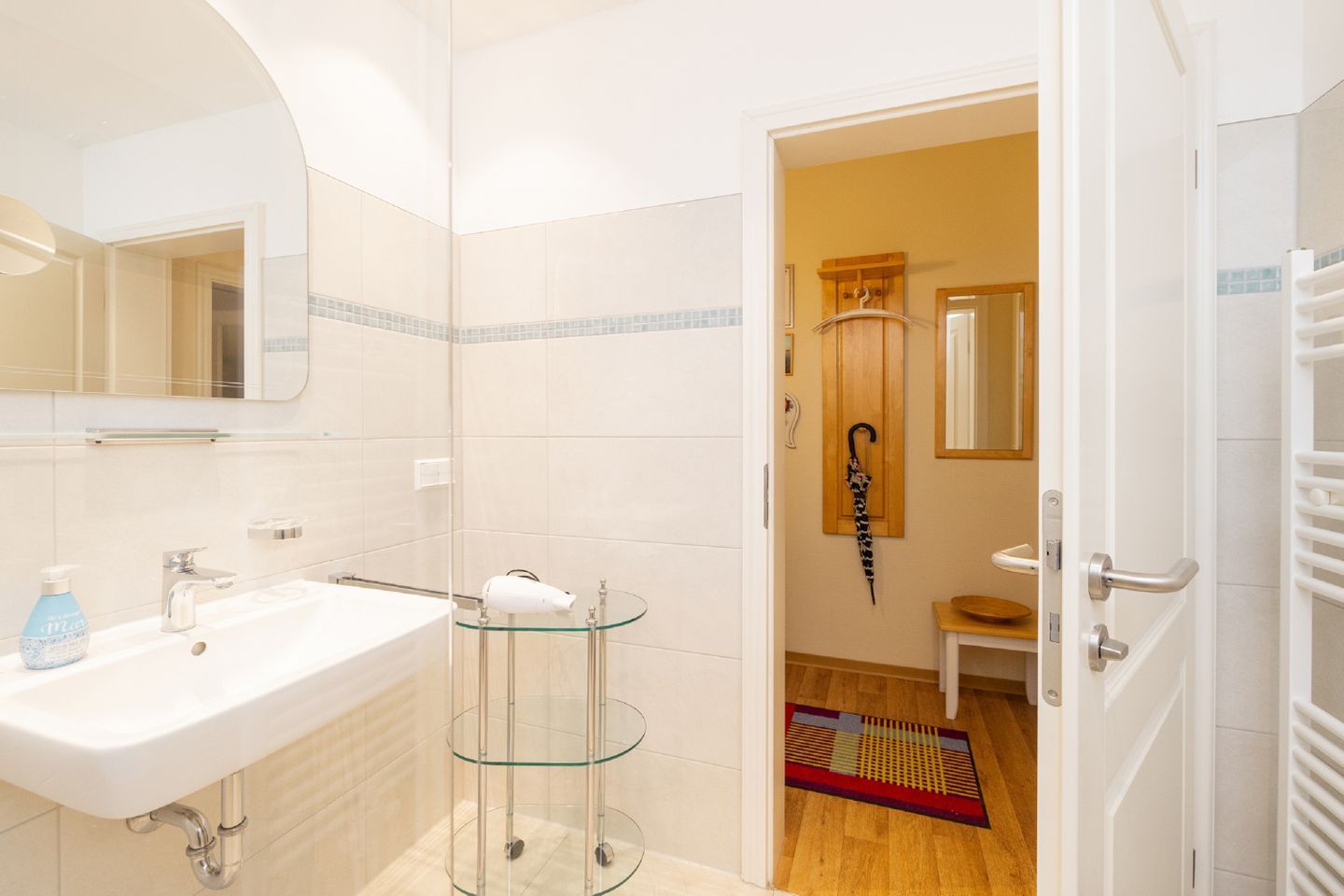 A modern bathroom with a white sink, glass shelves, a mirror, and a door leading to a hallway with a wooden floor and a patterned rug.